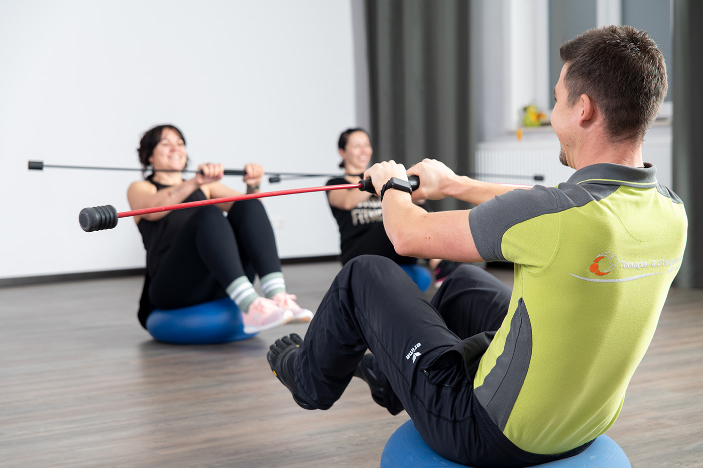 Eine Trainer und die Teilnehmer des individuellen Funktionstrainings sitzen auf Wackelkissen, während sie die Füße in der Luft halten und einen Wackelstab in den Händen halten.