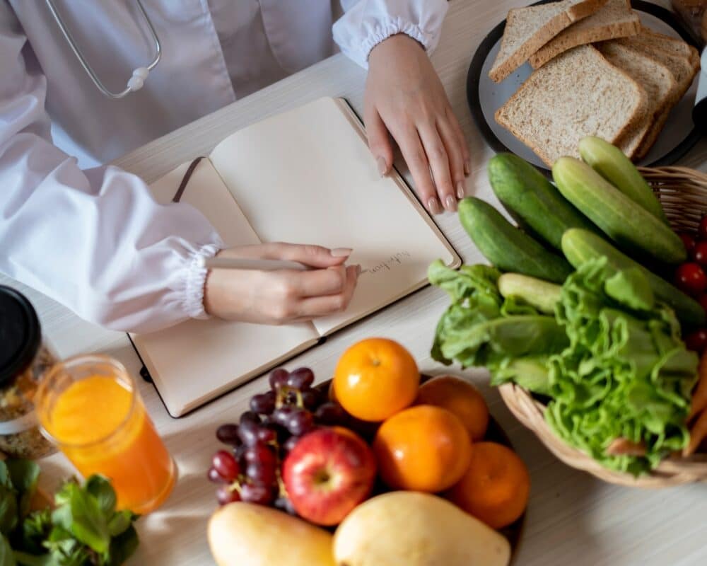 Arzt schreibt einen Plan zur Ernährungstherapie in ein Heft. Auf dem Tisch steht viel Obst und Gemüse.